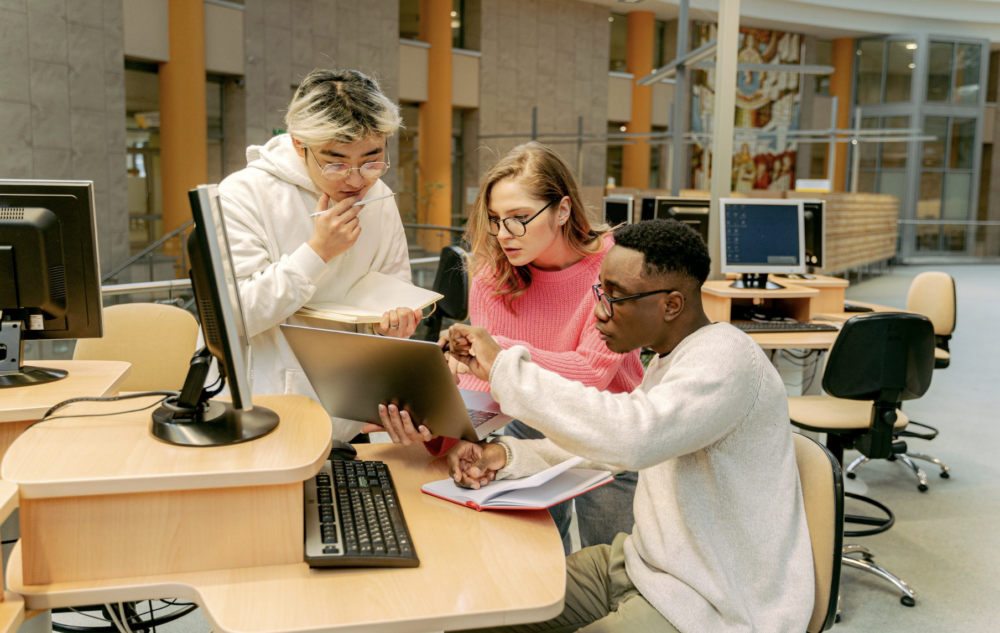 three individuals looking at a laptop screen. the environment is in a library.
